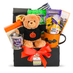A Halloween-themed gift basket featuring a plush teddy bear in a black sweater with a pumpkin face, an orange mug with a bat design, a tin of trick or treat, packaged snacks, and a black box tied with a bright orange ribbon and a 'Happy Halloween' tag.