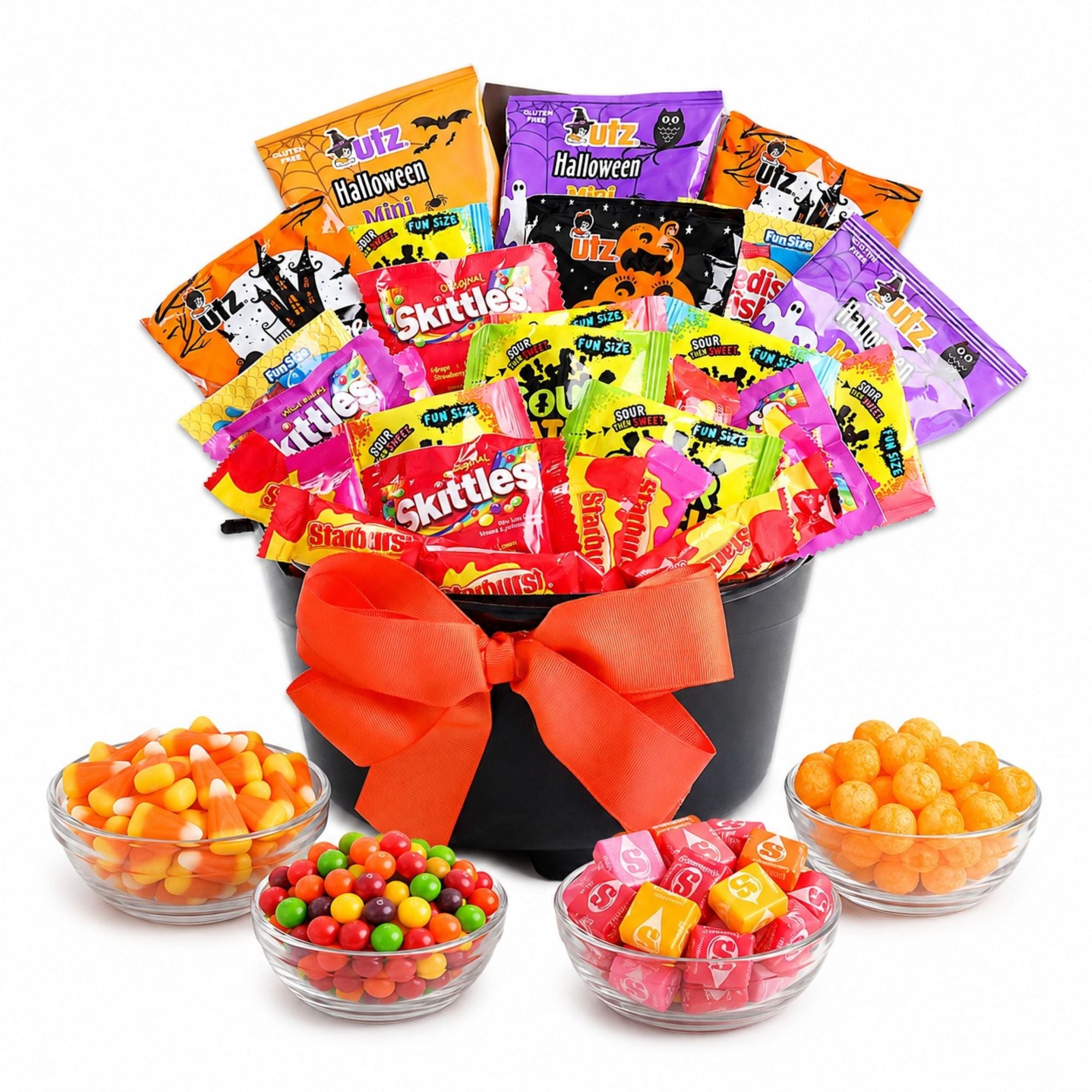 A Halloween-themed candy gift basket filled with assorted Skittles, and Starburst tied with a large orange bow in a black bucket, with four clear bowls of candy arranged in front on a white background.