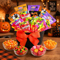 A Halloween-themed candy bouquet gift set in a black bucket with a large orange bow, filled with assorted candy bags. The bucket sits on a wooden table with bowls of candy and autumn decorations like pumpkins and a 'Trick or Treat' sign in the background.